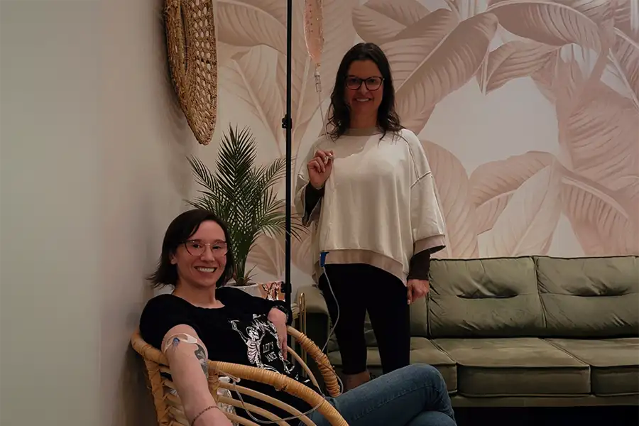 A professional at The Ivy Collective in Pawnee, IL standing beside a smiling client seated in a wicker chair receiving an IV therapy drip, in a warmly decorated room with a tropical leaf wallpaper mural.