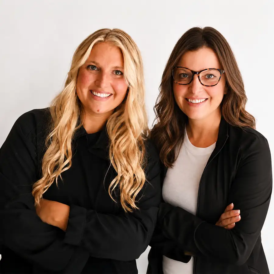 The two founders of The Ivy Collective in Pawnee, IL, mother-daughter duo Maddie Butcher and Jennifer Lambert, smiling with arms crossed in professional black attire against a white background.